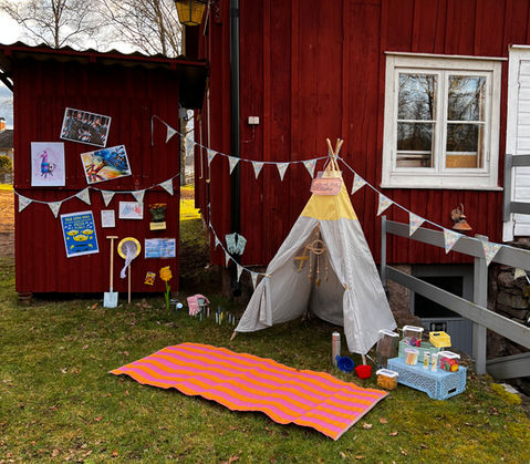 Ett tält med leksaker på en gräsmatta med en bygdegård i bakgrunden.