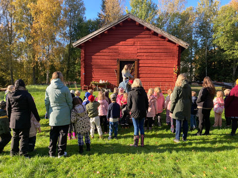 En grupp med barn och vuxna står utomhus i strålande sol och kollar på en föreställning av Teater Carpa.