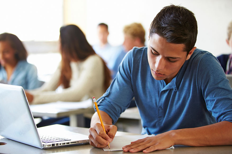 A young man is sitting in a classroom writing with a pencil on a sheet of paper. A laptop is open on the desk next to him. He is concentrating and looking at his writing.