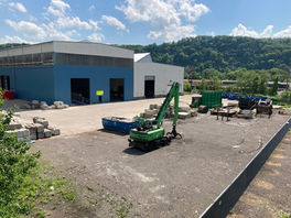 A blue and white warehouse sits on a large, fenced lot. Large concrete blocks are piled here and there. A green excavator with a grapple claw sits near an open dumpster.
