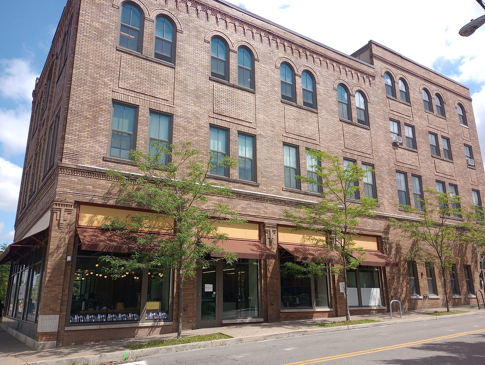 A three-story pinkish brick building has faded ochre awnings and mustard accents over large windows facing the street. Slender trees with pale green leaves grow every 20 feet or so in front of the windows.