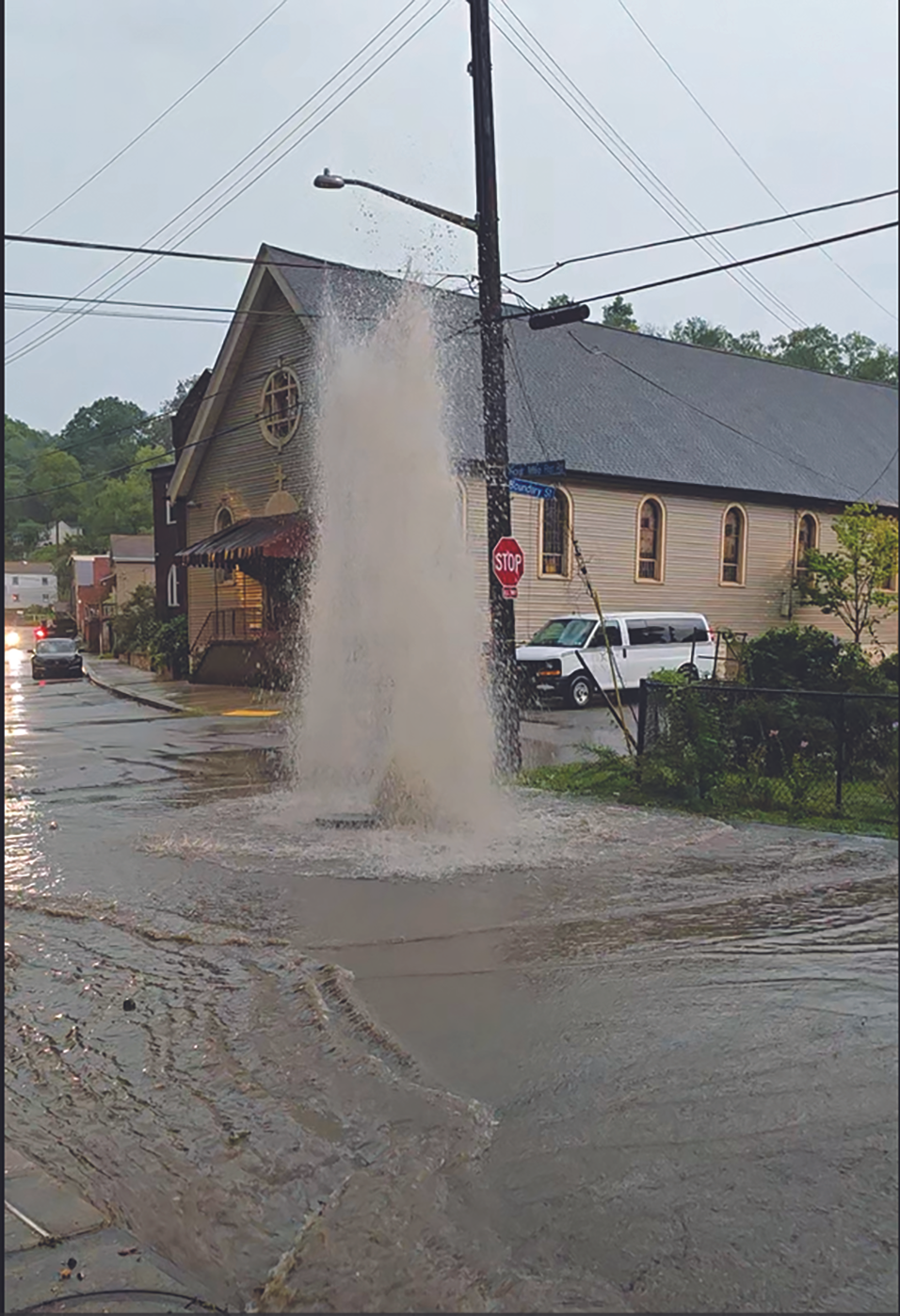 Brown water shoots up from a flooded intersection. A church and street signs are in the background.