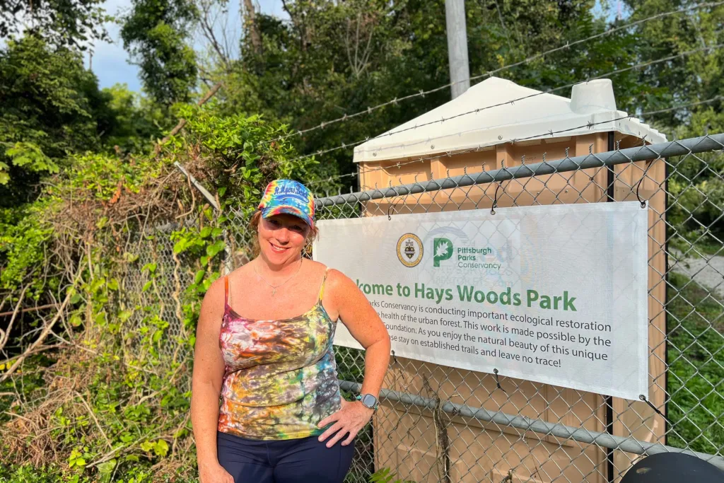 A woman in a tie-dye tank top and cap stands in front of a banner that reads, "Welcome to Hays Woods Park." 