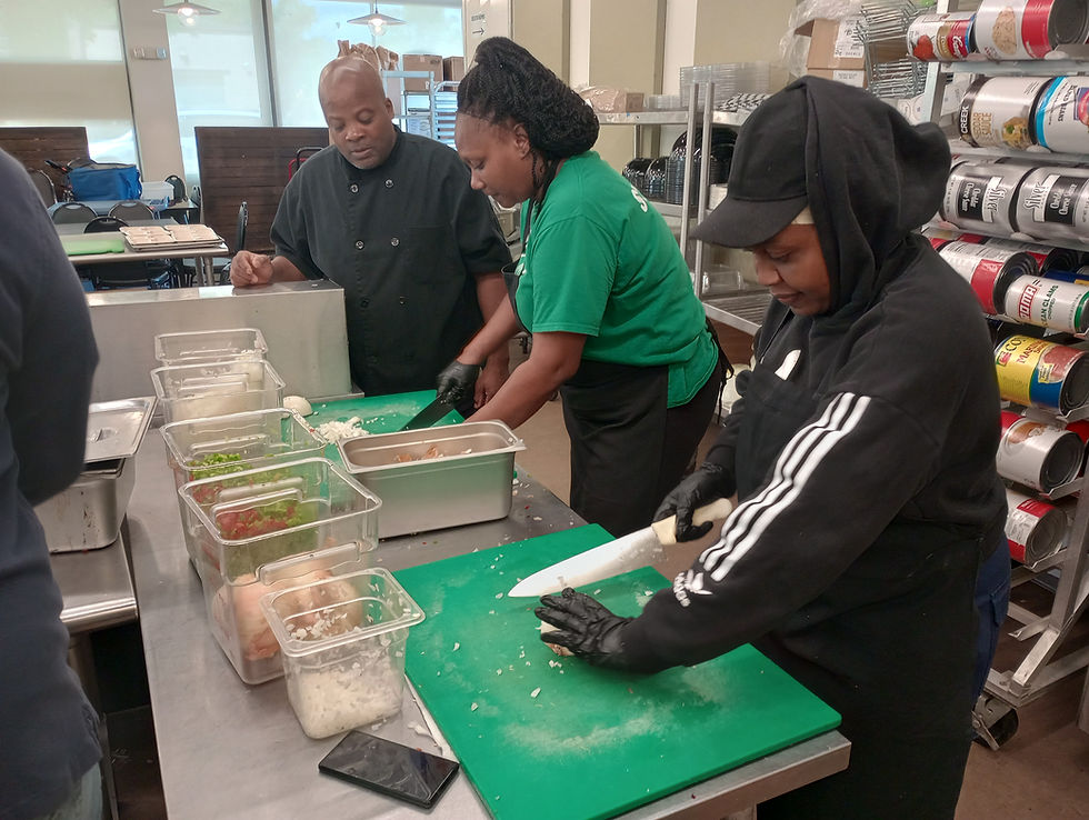 Two people in a kitchen chop veg and a person in a chef's uniform looks on.