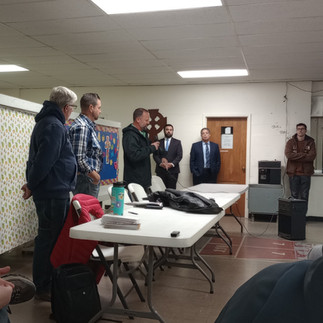 Six light-skinned men stand near white folding tables. The first three wear jeans with hoodies or flannel shirts. The other two wear suits. Another person stands on the far right side.