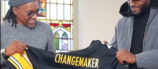 Two men stand holding a football jersey between them. The word "changemaker" is printed in gold on the jersey. 