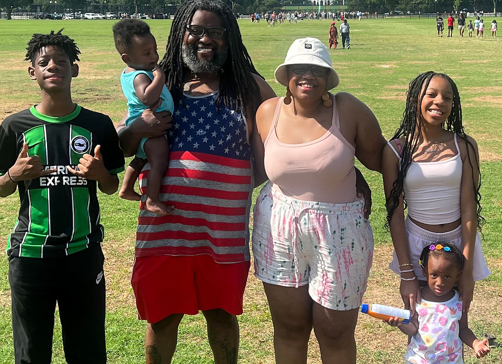 A family with two parents, two teens, a baby and a toddler stands in a field in the sun.