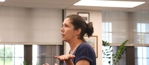 A light-skinned woman in a blue t-shirt and ponytail holds her hands up in disbelief or puzzlement.