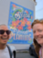 Large, colorful mural on a building reading “Trust the Seeds You Are Planting, Good Things Take Time,” decorated with pink and blue flowers, green leaves, and flowing decorative lettering on a bright blue background. Annelies (right) and Austin (left) smile in front of the mural looking into the camera.