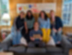 Five smiling people pose on a couch in a wood-paneled room under a Roots & Rivers banner; one holds a Roots Rivers plaque.