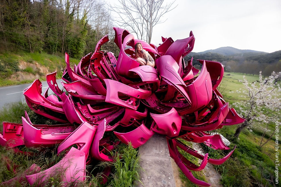 A vibrant outdoor sculpture made from vivid pink car parts, arranged in a blooming flower-like form, perched on an old stone wall. At the center, a woman—the artist Natalie Mandel ,nude and curled in a fetal position—emerges from the structure, gazing upward with a fragile, questioning expression. Set against a calm valley landscape with soft light and cloudy skies, the sculpture evokes both rebirth and motion.