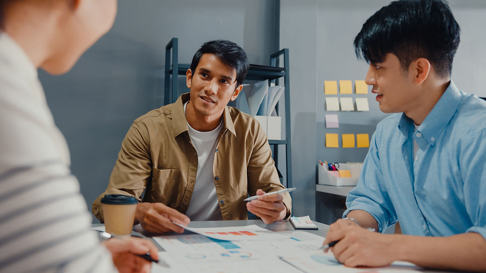 Three people discussing at a desk with papers and charts. Background has gray shelves and colorful sticky notes. Collaborative mood.