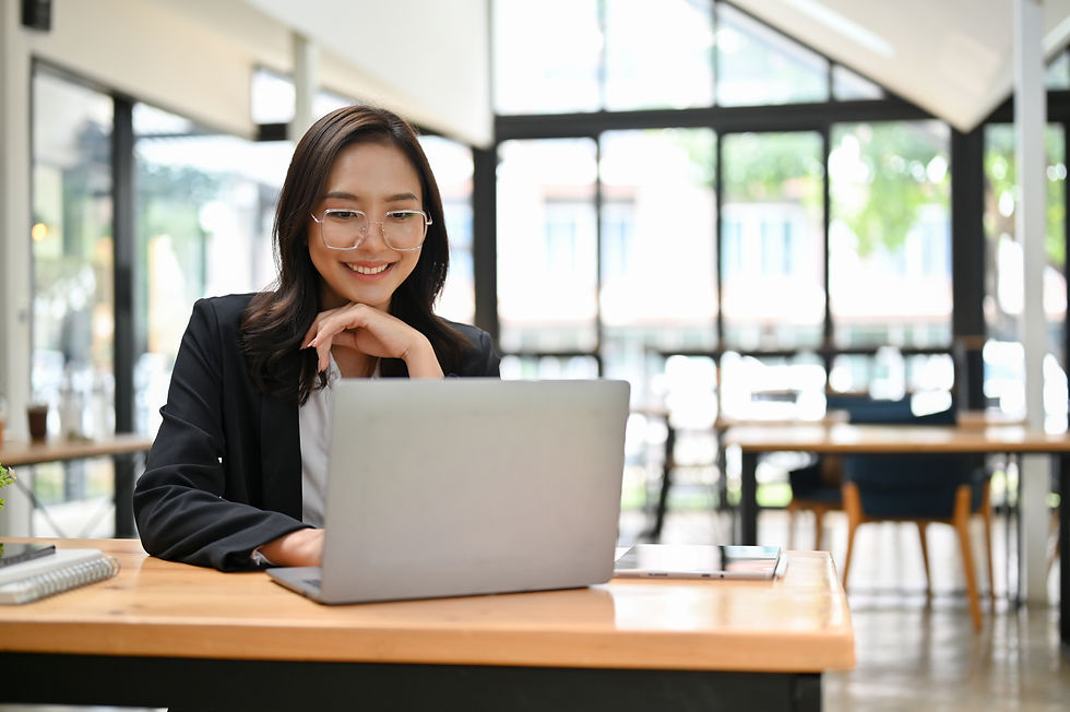 Smiling woman with glasses works on a laptop in a bright, modern office, seated at a wooden desk. Sunlight streams through large windows.