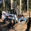 A man in carhartt overalls  is standing on the front of a white bobcat excavator. The excavator is in a forest with logs laying down around it.