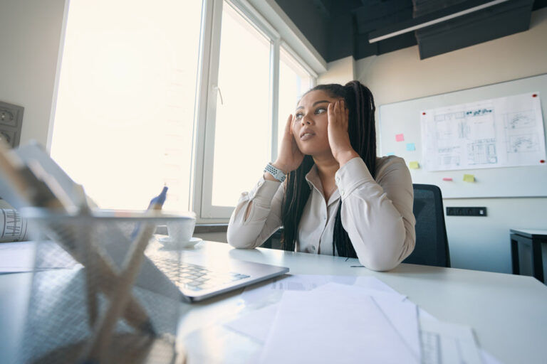 A woman sitting at a desk in an office, holding her head with both hands and staring upward, looking stressed or deep in thought.