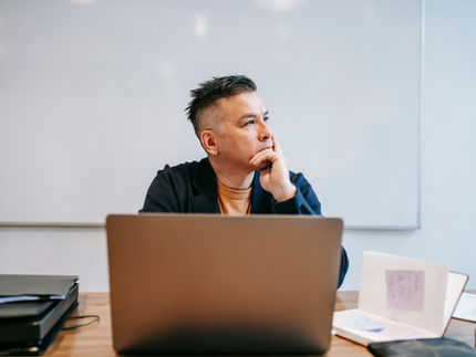 Man wearing an orange shirt and blue blazer sitting in an office with a laptop looking pensive