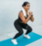 A woman wearing athletic clothing performs a squat exercise on a blue mat. Her expression is focused, and listening to music.