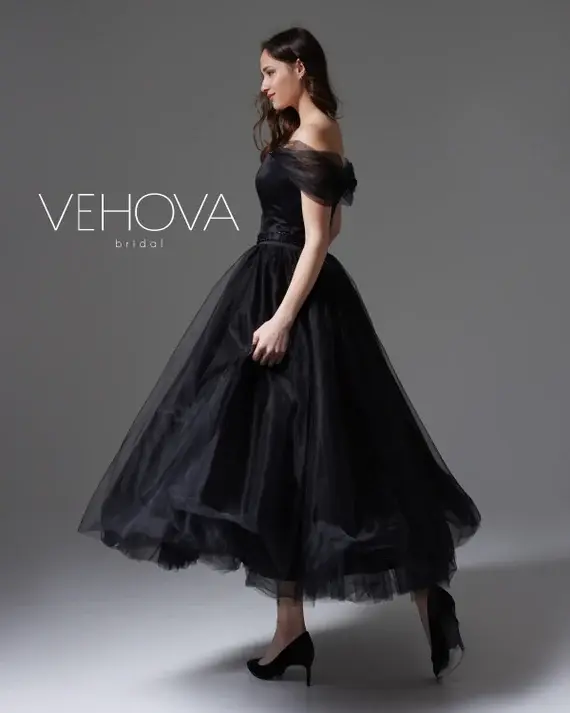 Woman in a flowing black off-shoulder gown poses confidently on gray backdrop. Text reads "VEHOVA bridal." Elegant and poised mood.