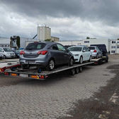 Laweta transportująca trzy samochody, w tym szary, na placu przemysłowym.