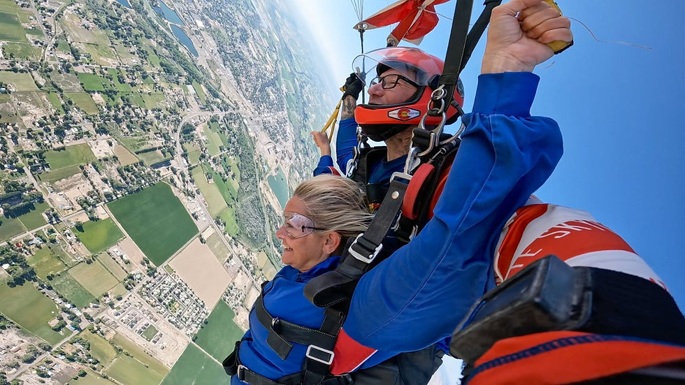 Tandem skydivers in blue suits and helmets soar above a patchwork landscape, one smiling, with a blue sky backdrop.