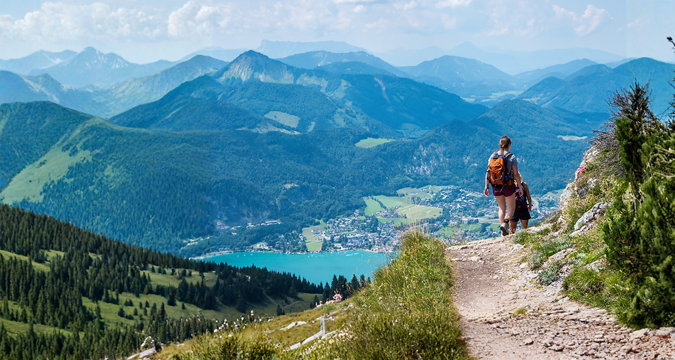 Hiker with orange backpack walks along a mountain trail overlooking lush valleys and a turquoise lake under a clear blue sky.