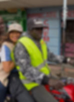 Two people ride a motorcycle through a street lined with small shops.