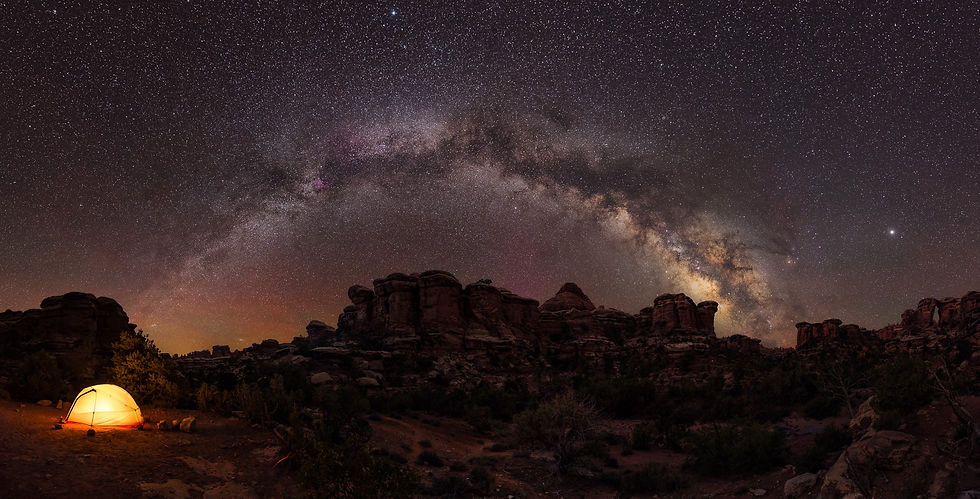 Under a breathtaking night sky filled with countless stars and the arching Milky Way, a lone tent glows warmly amidst the rugged rock formations of a remote desert landscape.