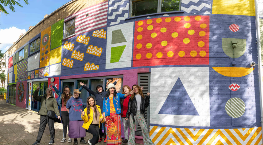 8 people are cheering and celebrating the completion of a Co-Creative Connection Community Mural with the Mayor of London and London catalyst on the Peabody community centre. Tower Hamlets, London.