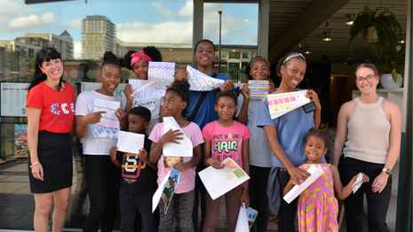 Group of smiling people holding colourful paper crafts in front of a building. Two adults on either side, including Rose, founder of CCC, and children in the middle. 