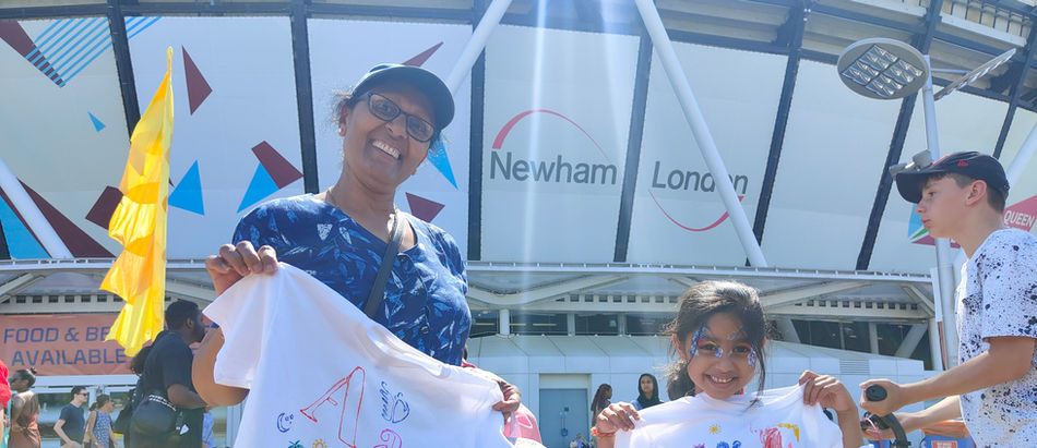 A adult and a child are standing outside West Ham football stadium on a sunny day. They are holding two t-shirts which have been drawn on, they are both smiling.