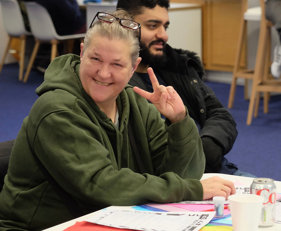 a woman is sitting a table smiling and holding up a peace sign for a workshop in partnership with Newham Council, Deafroots and SKILLS Community Mural Project, Newham 2023.