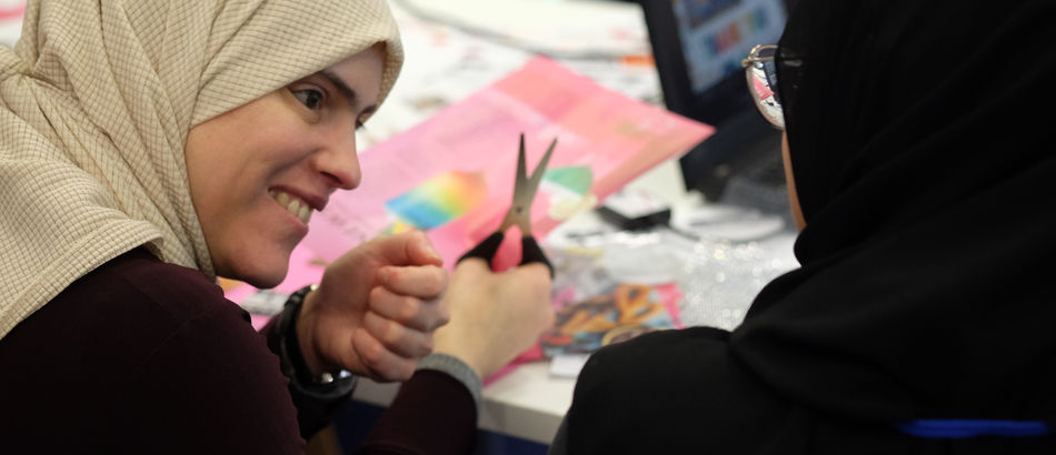 Two people are sitting at a desk, they are using scissors to cut out magazine clipping's.