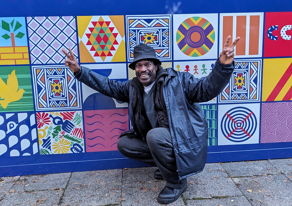 A man crouching in front of a colourful geometric CCC mural wall, with his arms raised in celebration. He's wearing a dark jacket and hat, smiling broadly. The mural behind him features various geometric patterns including diamonds, circles, waves, and other designs in bright colours including blues, reds, yellows, and greens.