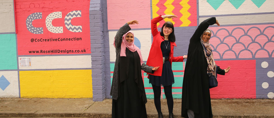 Three people are standing in front of a car park wall, they are creating the letter C with their arms. The wall has a freshly painted mural with pastel colours.