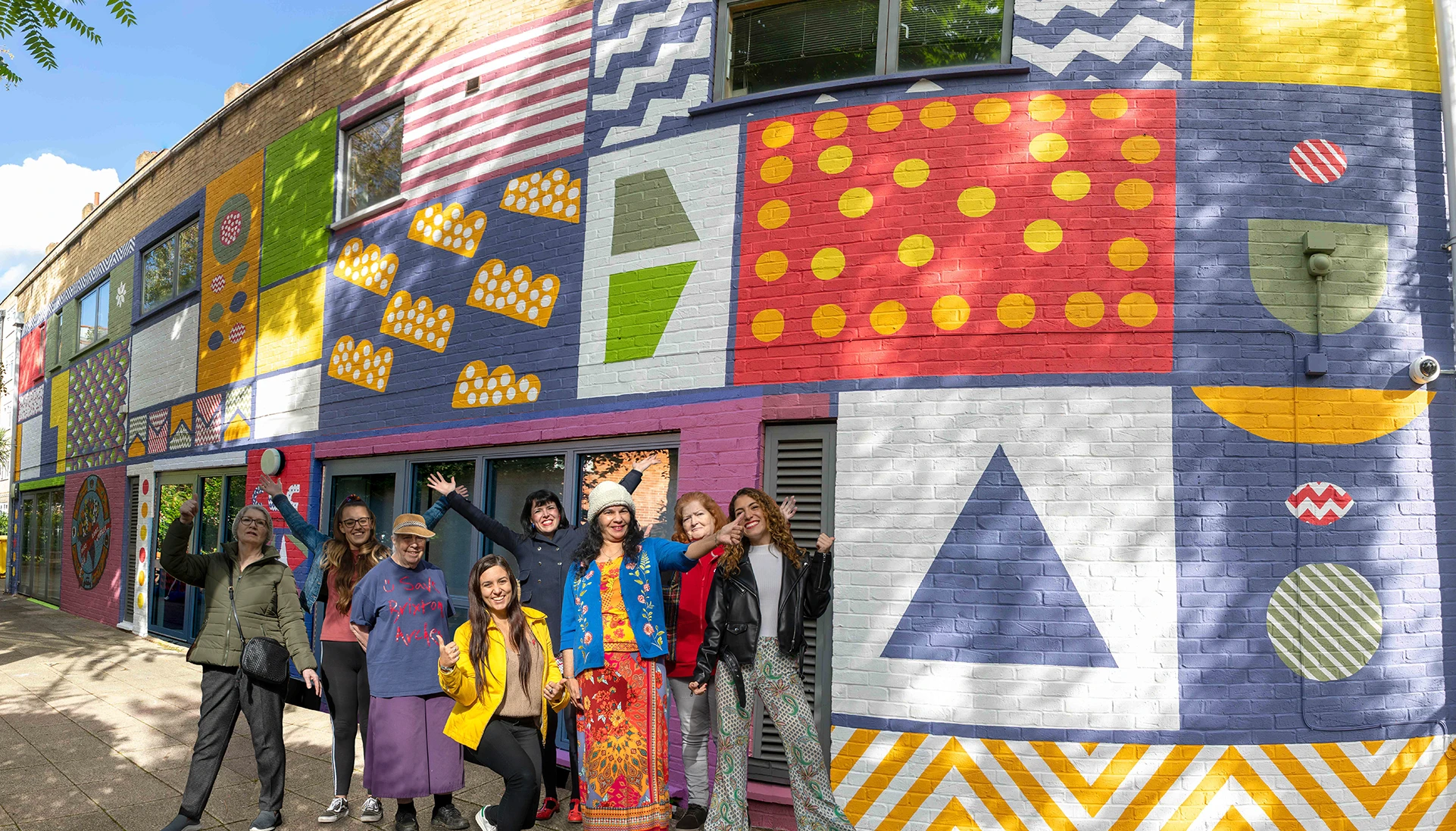 8 people are cheering and celebrating the completion of a Co-Creative Connection Community Mural with the Mayor of London and London catalyst on the Peabody community centre. Tower Hamlets, London.