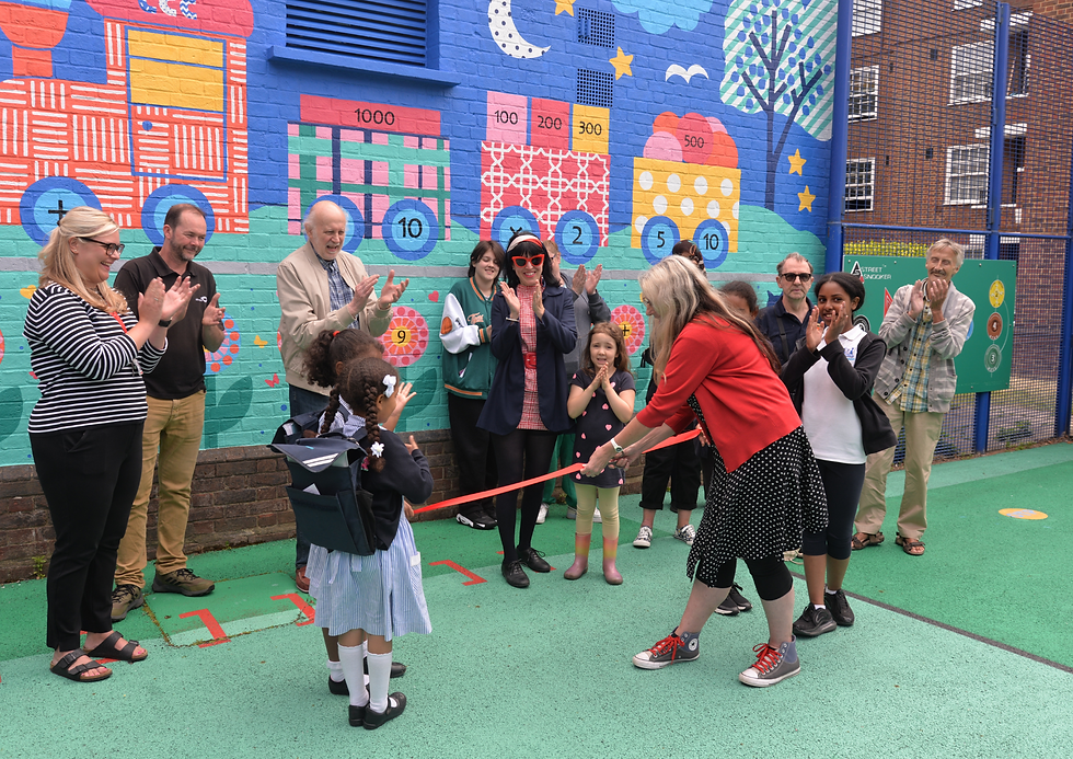 Ribbon-cutting unveiling for educational mural with colourful mathematical elements. Woman cutting ribbon while adults and kids stand in front of mural and clap/celebrate in school playground setting.