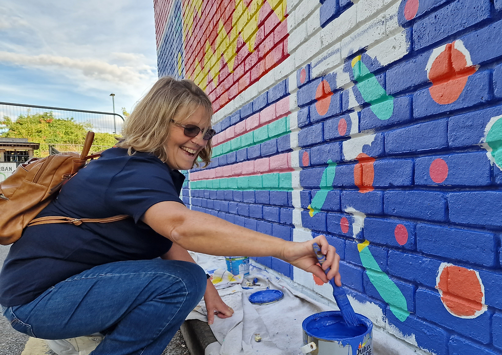 Smiling woman painting a colourful mural on a brick wall outdoors. She holds a paintbrush with blue paint, wearing glasses and a backpack.
