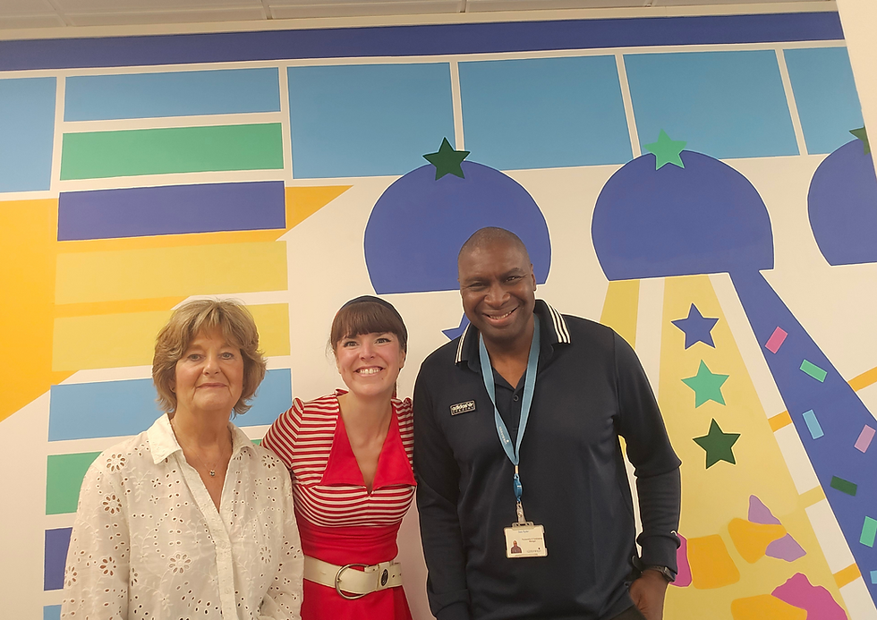 Rose, a woman and a man, smiling in front of a colourful geometric mural. Rose in red, the other woman in white, and the man in black with a name badge.