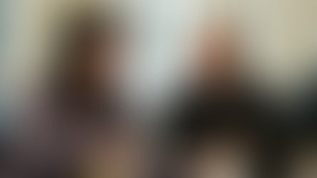 A woman and man sat at a table with art supplies and papers. The woman, with curly dark hair, is smiling while looking at papers she's holding. Beside her, the man, black jacket, is also smiling. The table has coloured markers, papers, mugs, and a vase with yellow flowers. 