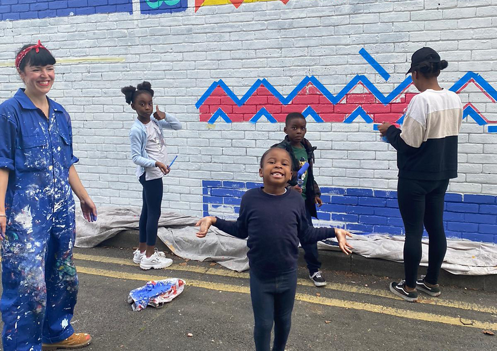 People painting a colourful mural on a brick wall. Blue, red zig-zag patterns. Two children look at the camera while one child smiles at the camera and poses. Rose watches and smiles. Playful, creative mood.