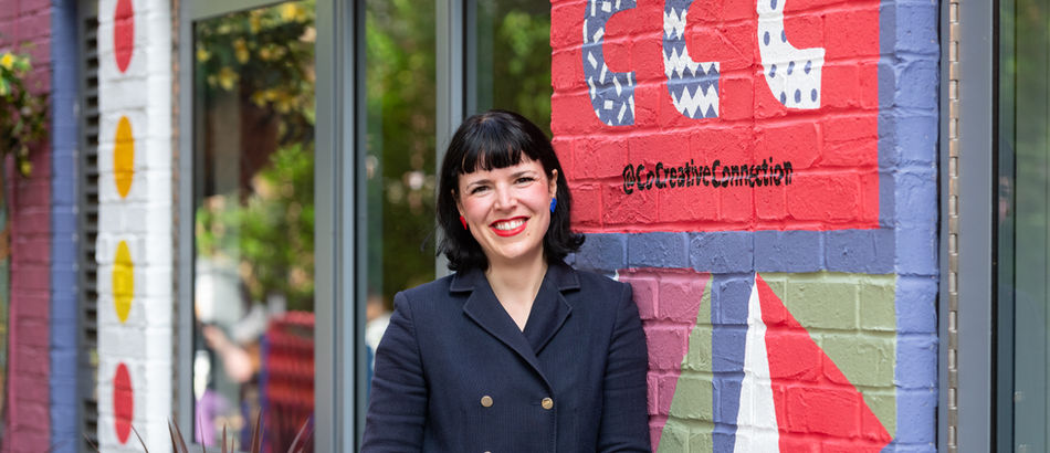 A lady with short black hair and a fringe is leaning against a wall. She is smiling and wearing a double breasted navy coat. The wall has geometric patterns painted on,