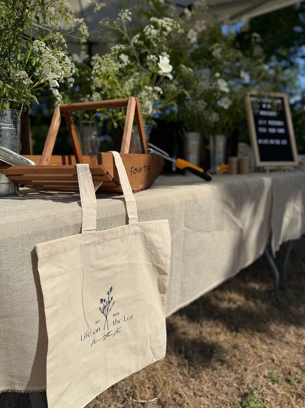 Canvas tote bag on a table with flower motif, surrounded by wildflowers, wooden tray, and gardening tools. Sunny outdoor market scene.