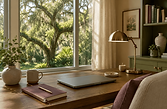 Warm Central Florida home office with a wooden desk, closed laptop, mauve notebook, brass lamp, ceramic mug, and live oak trees visible through the window.