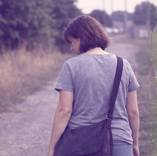Documentary-style photograph of an adult walking alone along a quiet outdoor path, captured candidly from behind in soft natural daylight, conveying a private, reflective moment without posed expression or destination focus.