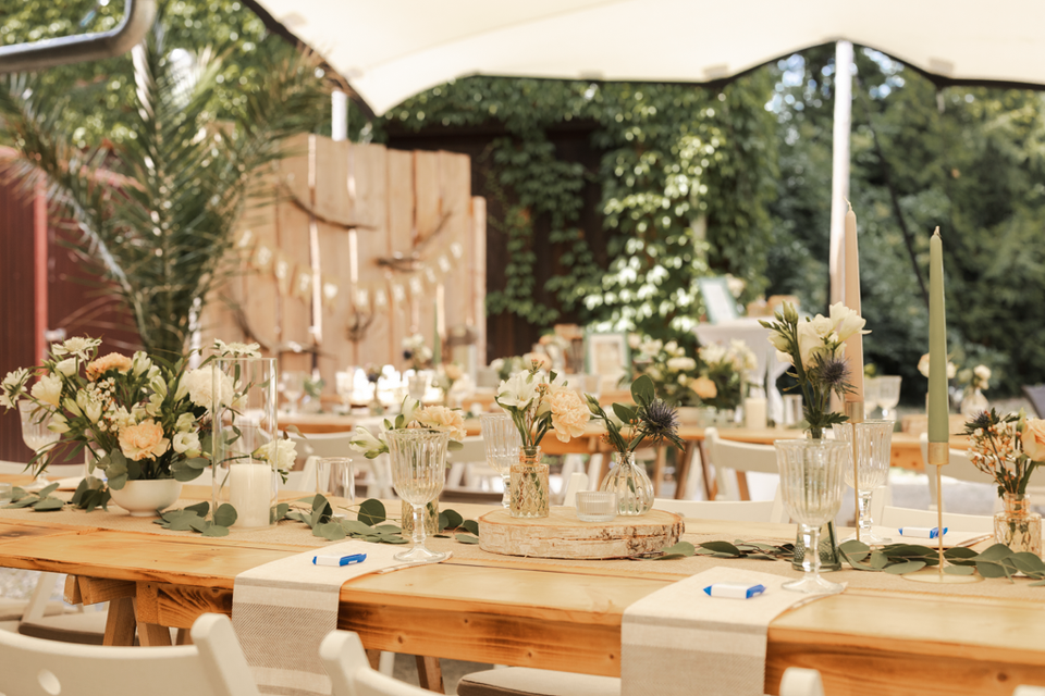 Festlich gedeckte lange Holztische unter einer weißen Stretchtent im Garten des Gasthofs Mügeln bei Jessen. Die Tische sind dekoriert mit frischen Blumenarrangements in Vasen, Kerzen, Weingläsern und cremefarbenen Tischläufern. Im Hintergrund sind weitere Tische, grüne Pflanzen und rustikale Holzelemente zu sehen. Eine einladende Atmosphäre für Hochzeiten.