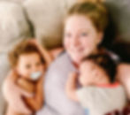 A woman is smiling on a couch, holding a sleeping baby and a toddler with a pacifier, creating a warm, affectionate mood.