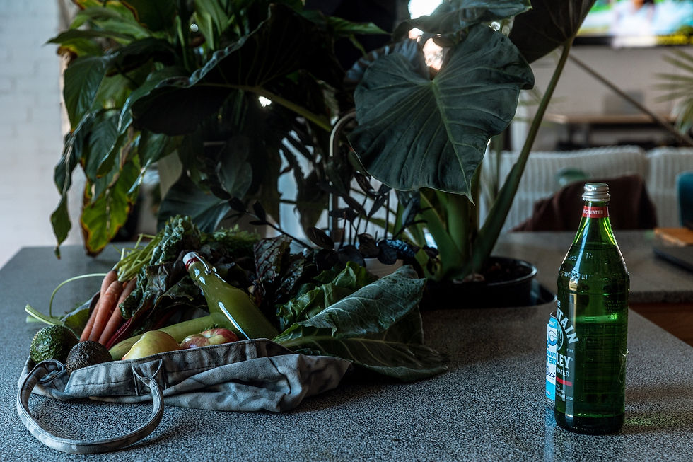 Fresh produce and a green juice bottle sit in a bag on a table near lush green plants. A green glass bottle is nearby. Cozy indoor setting.