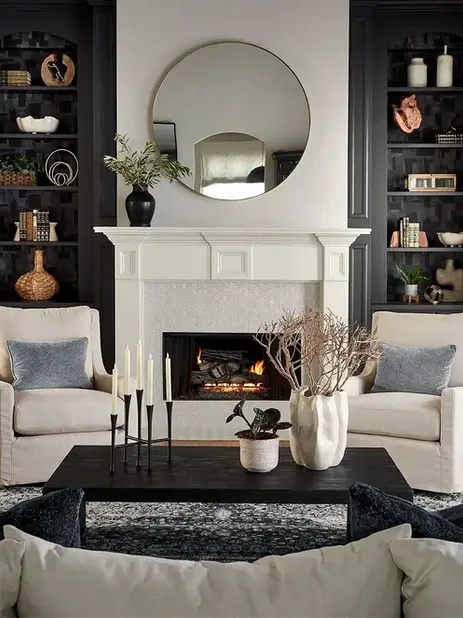 2-story transitional living room | family room | large white sofa couch | white upholstered swivel arm chair | white herringbone tile fireplace surround | urbane bronze | custom builtins | builtin bookcase | black coffee table | neutral beige curtains | round fireplace mirror | blue area rug | arched doorway | blue abstract wall art paintings | metal brass dome pendant light