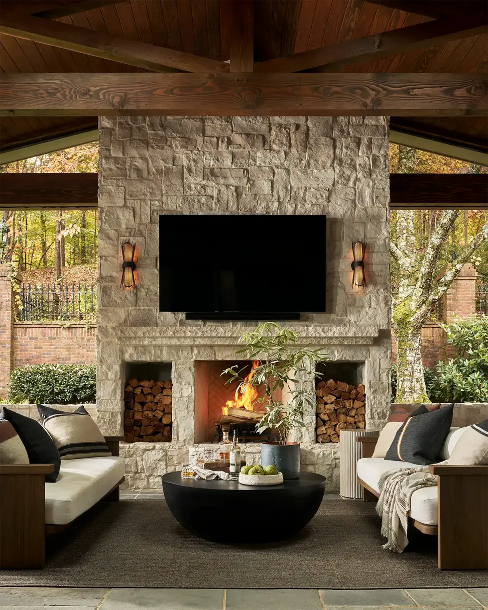 Floor-to-ceiling natural stone outdoor fireplace with firewood storage niches, mounted television, and sculptural sconces