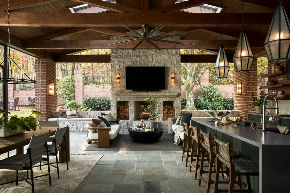 Covered outdoor entertaining pavilion with kitchen, dining table, and fireside seating beneath vaulted timber trusses and skylights